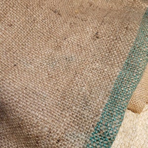 May include: Close-up of a textured burlap fabric with a natural brown color. A teal-colored stripe runs vertically along the right side. The coarse weave of the material is visible, suggesting a rustic or agricultural theme.