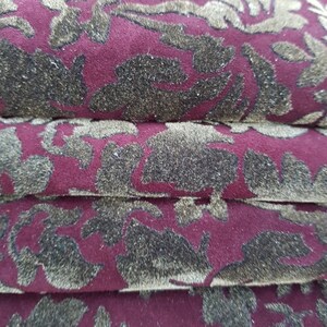 May include: Close-up of a burgundy fabric with a gold floral pattern. The fabric is folded in layers, showing the texture and detail of the design.