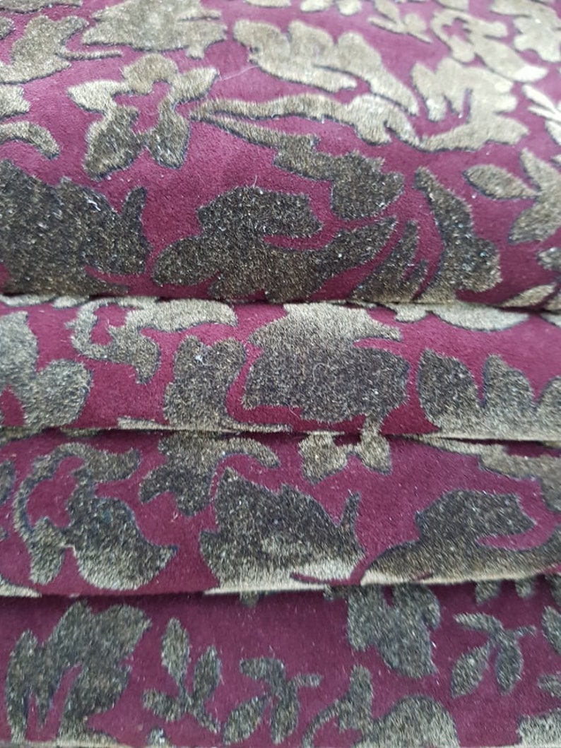 May include: Close-up of a burgundy fabric with a gold floral pattern. The fabric is folded in layers, showing the texture and detail of the design.