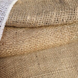 May include: Close-up of a stack of burlap fabric. The coarse, woven material is a natural tan color, with a white mesh trim visible on the left side. The texture of the burlap is prominent, showing the individual fibers.