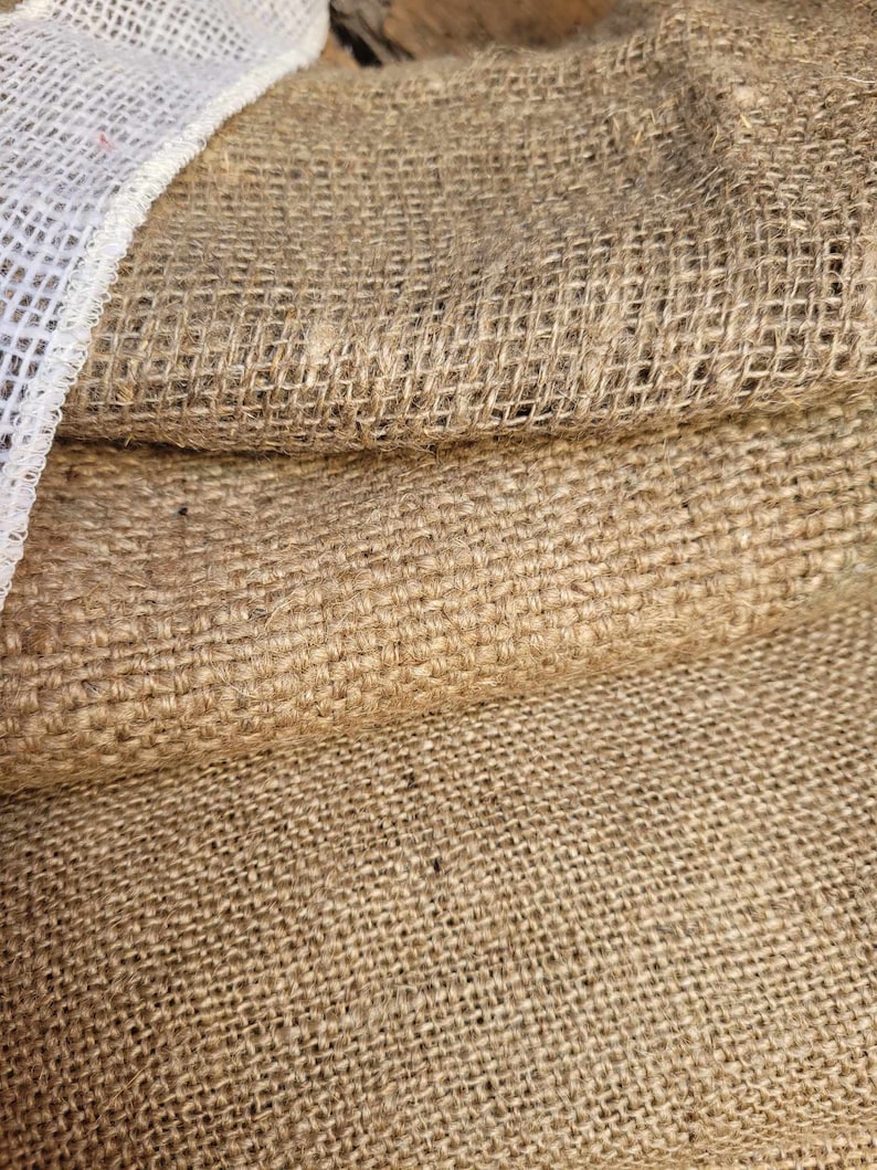 May include: Close-up of a stack of burlap fabric. The coarse, woven material is a natural tan color, with a white mesh trim visible on the left side. The texture of the burlap is prominent, showing the individual fibers.
