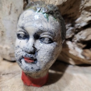 May include: A ceramic sculpture of a human head with a cracked, off-white surface. The face has black details, red lips, and a red base. The hair has green accents.