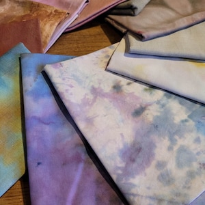 Op de afbeelding: Een verzameling stoffen vierkanten in verschillende tinten bruin, paars, blauw en grijs. De stof heeft een tie-dye patroon en is in een cirkelvormige opstelling op een houten oppervlak geplaatst.