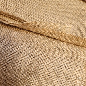 May include: Close-up of a roll of natural burlap fabric. The coarse weave of the tan-colored material is visible, with the fabric folded over itself. The texture suggests a rustic or farmhouse aesthetic, suitable for crafts or decor.