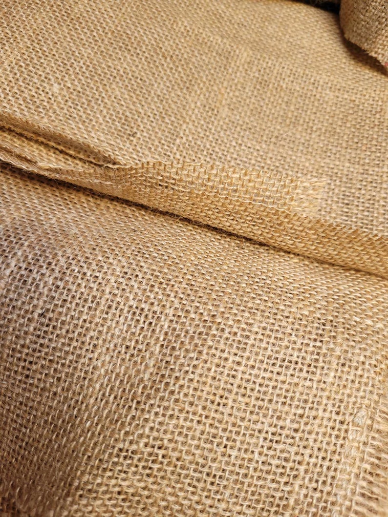 May include: Close-up of a roll of natural burlap fabric. The coarse weave of the tan-colored material is visible, with the fabric folded over itself. The texture suggests a rustic or farmhouse aesthetic, suitable for crafts or decor.