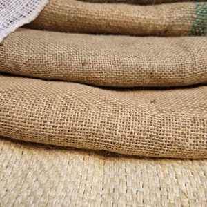 May include: Close-up of several layers of burlap fabric. The fabric is a natural tan color with a coarse weave. A white mesh fabric is visible on top of the burlap. The image highlights the texture and natural tones of the material.