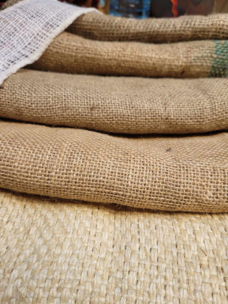 May include: Close-up of several layers of burlap fabric. The fabric is a natural tan color with a coarse weave. A white mesh fabric is visible on top of the burlap. The image highlights the texture and natural tones of the material.