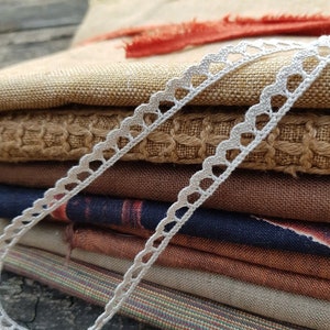 Puede incluir: Una pila de muestras de tela en varios colores y texturas, incluyendo marr&oacute;n, beige y azul, con un ribete de encaje blanco tejido a crochet.
