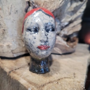 Puede incluir: Una escultura de cerámica de una cabeza femenina con cabello rojo y labios rojos. La cara tiene una apariencia agrietada y envejecida con motas negras. La cabeza está montada sobre una base oscura y brillante.