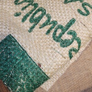 May include: Close-up of a burlap sack with green embroidered text. The text appears to be a brand name or product description. The burlap has a natural, woven texture, with a green border.