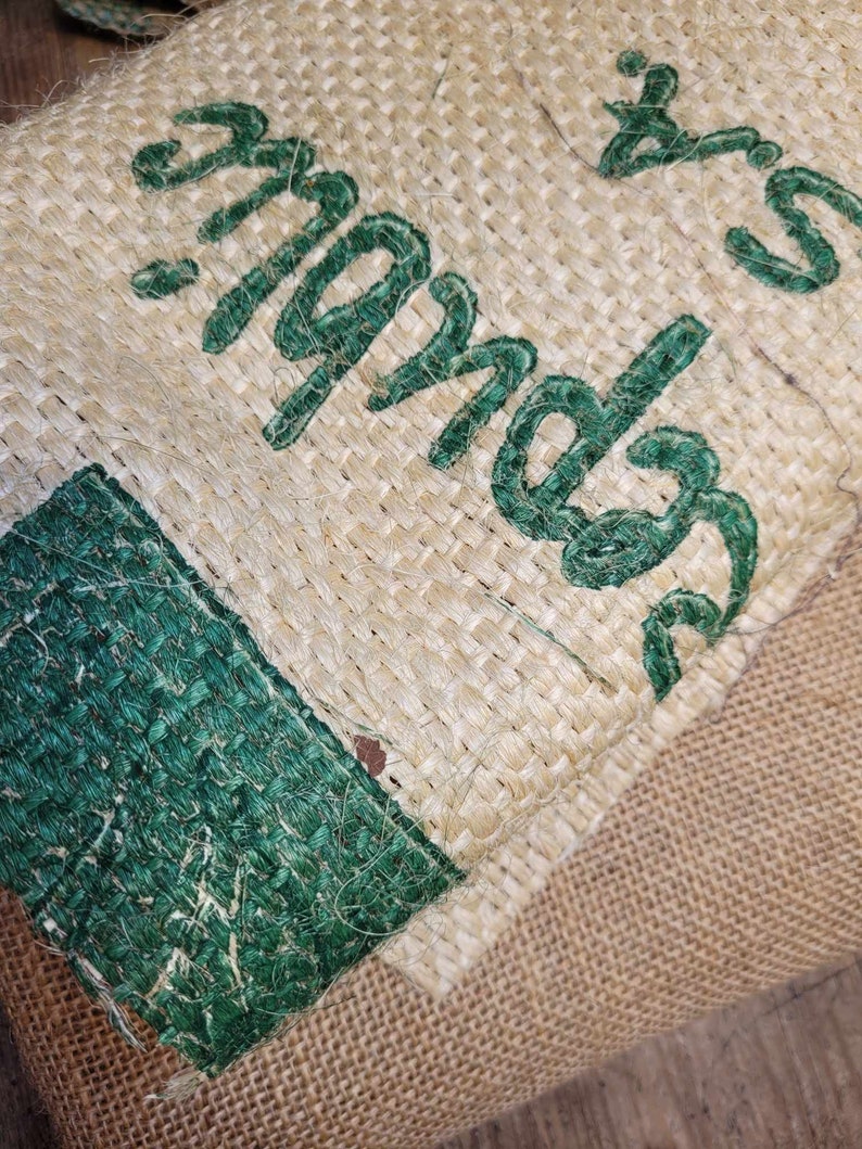 May include: Close-up of a burlap sack with green embroidered text. The text appears to be a brand name or product description. The burlap has a natural, woven texture, with a green border.