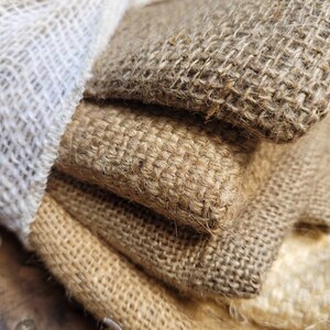 May include: Close-up of stacked burlap fabric in various shades of brown and tan. A white, loosely woven fabric is visible on the left side. The coarse texture of the burlap is prominent, with visible fibers and a natural, rustic appearance.