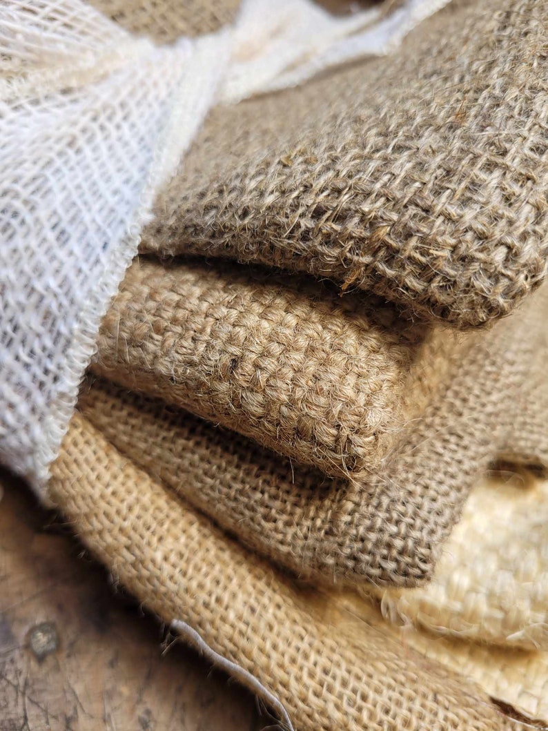 May include: Close-up of stacked burlap fabric in various shades of brown and tan. A white, loosely woven fabric is visible on the left side. The coarse texture of the burlap is prominent, with visible fibers and a natural, rustic appearance.