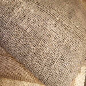 May include: Close-up of several layers of burlap fabric in varying shades of brown and tan. The fabric has a coarse, open weave, with visible texture and a natural, rustic appearance. The layers are folded and stacked, showcasing the different tones.