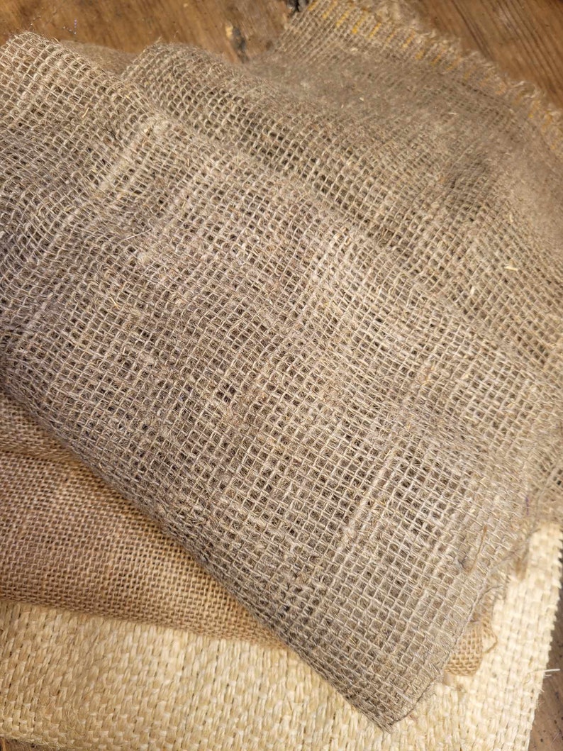 May include: Close-up of several layers of burlap fabric in varying shades of brown and tan. The fabric has a coarse, open weave, with visible texture and a natural, rustic appearance. The layers are folded and stacked, showcasing the different tones.