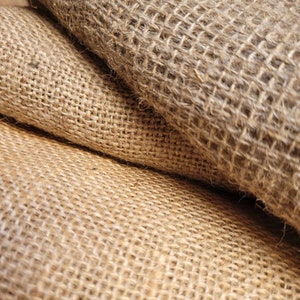 May include: Close-up of rolled burlap fabric. The natural fiber material has a coarse, open weave, with shades of tan and brown. The texture is rough and the fabric appears to be folded over itself, creating depth and shadow.