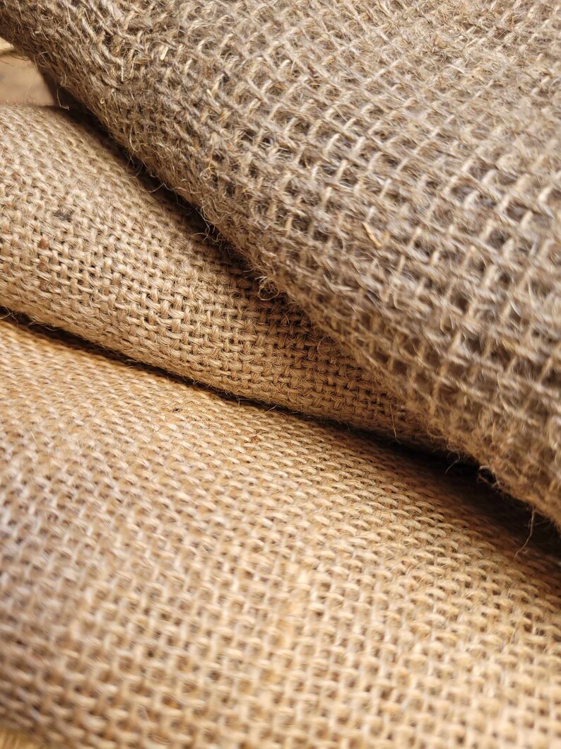 May include: Close-up of rolled burlap fabric. The natural fiber material has a coarse, open weave, with shades of tan and brown. The texture is rough and the fabric appears to be folded over itself, creating depth and shadow.
