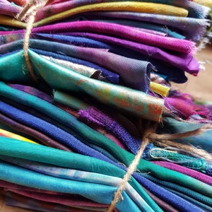 Könnte beinhalten: Ein Bündel bunter Stoffreste, mit Bindfaden zusammengebunden. Die Stoffstücke zeigen eine Vielzahl von Farben, darunter Schattierungen von Lila, Blau, Grün und Rosa. Der Stoff scheint Seide oder ein ähnliches Material zu sein, mit leichtem Glanz.