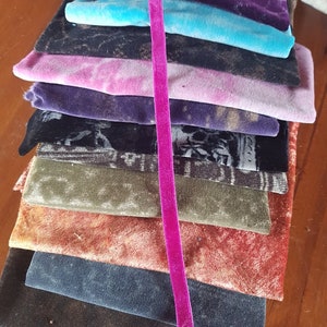 May include: A stack of fabric swatches in various colours and textures, including black, blue, green, orange, and purple. A pink velvet ribbon runs vertically down the centre of the stack.