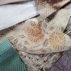 May include: A selection of fabric samples in diverse textures and colours. Features white crocodile-effect leather, floral embossed fabric, and shimmering teal and silver materials. The swatches are arranged in a layered composition.
