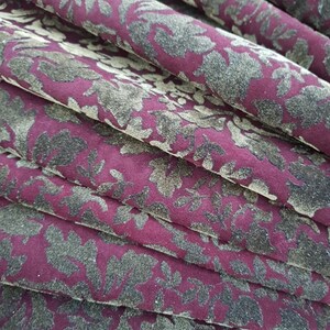 May include: Close-up of a burgundy fabric with a repeating pattern of dark green floral designs. The fabric is folded in layers, creating a textured effect.