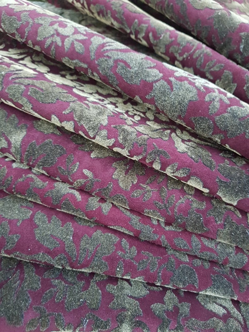 May include: Close-up of a burgundy fabric with a repeating pattern of dark green floral designs. The fabric is folded in layers, creating a textured effect.