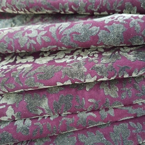 May include: Close-up of a burgundy fabric with a gold floral pattern. The fabric is folded in multiple layers, creating a textured effect.