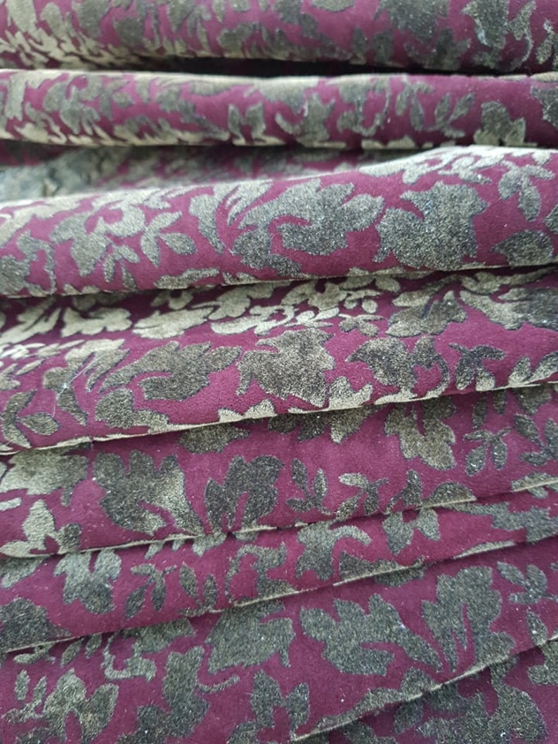 May include: Close-up of a burgundy fabric with a gold floral pattern. The fabric is folded in multiple layers, creating a textured effect.