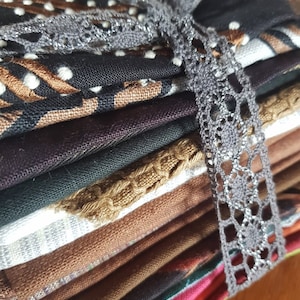Könnte beinhalten: Ein Stapel Stoffquadrate in verschiedenen Farben und Mustern, gebunden mit einem grauen Spitzenband. Die Stoffe umfassen Brauntöne, Schwarz, Grün und Rot, mit Designs wie Tupfen und geometrischen Formen. Das Band sorgt für eine dekorative Note.