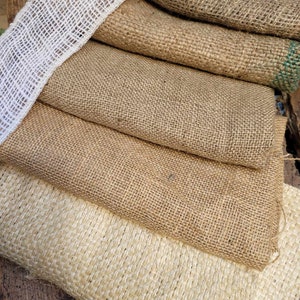 May include: A stack of burlap fabric in various shades of brown and beige. A white mesh ribbon is draped over the top. The coarse texture of the fabric is visible, suggesting a rustic or natural aesthetic. The image showcases the different textures and colors of the burlap.