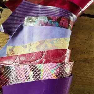 May include: A collection of colorful leather swatches in shades of purple, pink, and gold. The textures vary from smooth to embossed, including a snakeskin pattern. The swatches are arranged in a fan shape, showcasing the different materials.