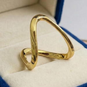 Puede incluir: Un anillo dorado con un diseño abierto y singular, presentado en una caja de anillo de terciopelo azul con interior crema. El anillo tiene una forma abstracta y moderna con líneas suaves y curvas.