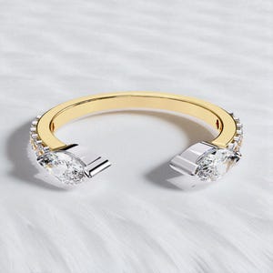 Puede incluir: Anillo de brazalete abierto con una banda dorada y dos extremos plateados, cada uno con una gema grande y transparente con corte marquesa. La banda está bordeada con pequeñas gemas transparentes.