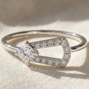 Könnte beinhalten: Ein silberner Ring mit einem quadratisch geschliffenen Diamanten und einem dekorativen Band, das mit kleinen Diamanten besetzt ist. Das Design des Rings ist elegant und betont das Funkeln der Edelsteine.
