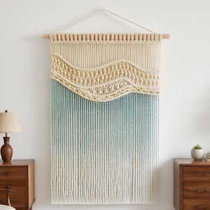 Op de afbeelding: Een grote, handgemaakte macramé wandhanger. Het bovenste gedeelte is crèmekleurig met een geweven ontwerp, dat overgaat in een lichtblauwe en witte gradiënt. Het hangt aan een houten roede met een natuurlijke vezelkoord.