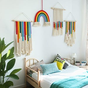 May include: A collection of handmade macrame wall hangings in a bedroom setting. The pieces feature rainbow-colored yarn and off-white fringe. One is a rainbow shape, and the others are rectangular with colorful vertical stripes.