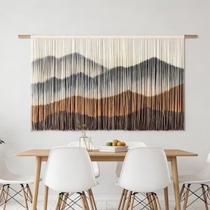 May include: A large wall hanging with a mountain landscape design. The artwork features a gradient of colors, transitioning from cream at the top to shades of gray and brown at the bottom. It is mounted on a wooden rod above a dining table.