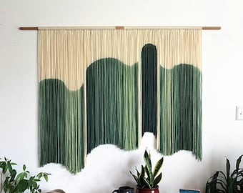 May include: A wall hanging made of yarn with a green and white ombre design. The hanging is made of many strands of yarn that are tied together to create a wave-like pattern. The hanging is mounted on a wooden dowel rod.