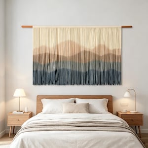 May include: A large, rectangular wall hanging with a mountain-like design. The top is cream-colored, transitioning to brown and then to a deep blue at the bottom. It is suspended from a wooden rod, creating a textured, layered effect.