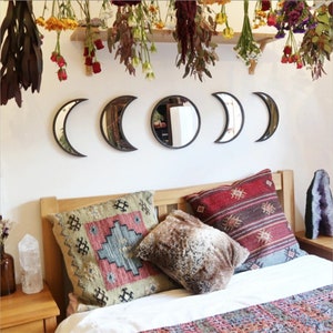 May include: Five crescent moon-shaped mirrors with black frames are arranged on a white wall. The mirrors are hung above a bed with a wooden headboard and colorful patterned pillows.