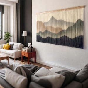 以下が含まれることがあります： 山岳風景のデザインが施された、長方形の大きな壁掛け。上部はベージュ、下部は濃い青色で、その間に緑と茶色の色合いがグラデーションで表現されています。天然木のフレームに取り付けられています。