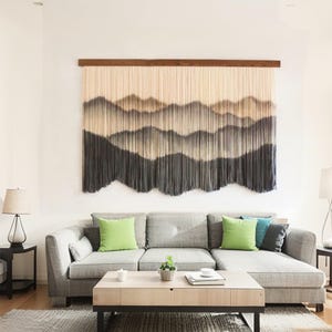 Könnte beinhalten: Große, strukturierte Wandbehänge, die eine Bergkette in Grau- und Cremetönen darstellen. Das Kunstwerk ist an einer Holzstange über einem grauen Ecksofa mit grünen und blauen Zierkissen befestigt. Eine moderne Wohnzimmereinrichtung.