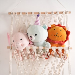 May include: A cream macrame shelf displaying three soft toys: a pink elephant, a light blue bear with a party hat, and an orange lion. The shelf is made of knotted rope and wooden dowels, suitable for a nursery or children's room.