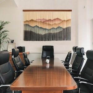 Könnte beinhalten: Ein rechteckiger Wandbehang mit einem Berglandschaftsdesign in Beige-, Braun- und Grautönen. Das Kunstwerk ist auf einem Holzrahmen montiert und hängt über einem langen Konferenztisch aus Holz, umgeben von schwarzen Lederstühlen.