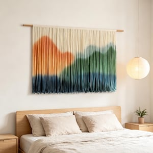 Peut inclure: Une tenture murale composée de brins de laine verticaux couleur crème avec un motif de paysage teint par immersion. Le design présente une grande forme de montagne orange à gauche, passant par des sommets verts et une base bleu foncé, suspendue à une tige en bois.