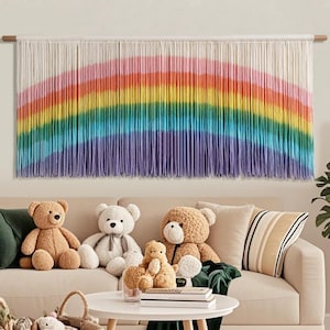 May include: A vibrant rainbow-coloured macrame wall hanging, with a spectrum of pink, orange, yellow, green, blue, and purple. Beneath, a beige sofa is decorated with teddy bears and cushions, creating a warm and inviting space.