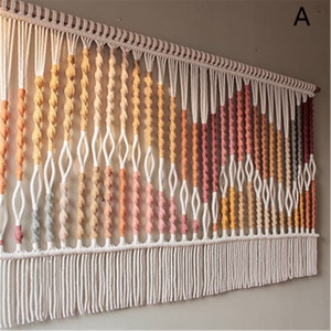Op de afbeelding: Macramé wandkleed met een kleuroverloop van perzik, oranje en roze. Het ontwerp heeft geknoopte secties en een franje aan de onderkant. De letter "A" staat in de rechterbovenhoek.