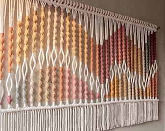 May include: Macrame wall hanging with a gradient of colors, including peach, orange, and pink. The design features knotted sections and a fringe at the bottom. The letter "A" is in the top right corner.
