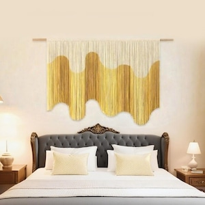 Peut inclure: Une tenture murale tissée avec un motif ondulé dans des tons or et crème. L'œuvre d'art textile est fixée sur une tringle en bois et suspendue au-dessus d'un lit avec une tête de lit grise et une literie blanche. La pièce est décorée dans des tons neutres.