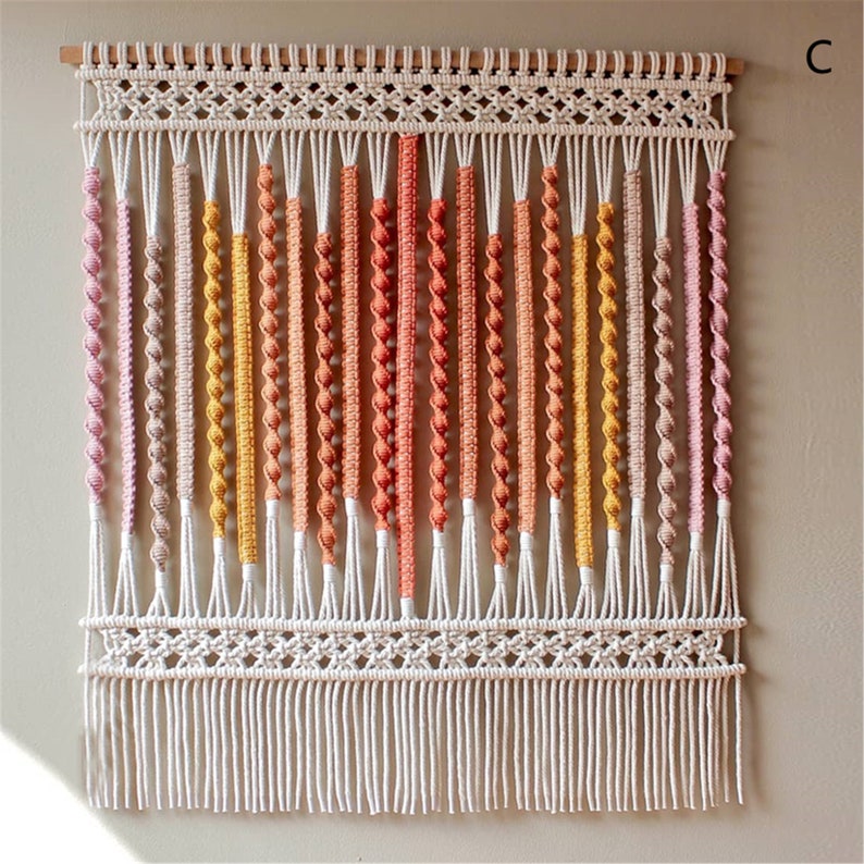May include: Macrame wall hanging with a wooden dowel and a cream-colored woven top. Vertical strands of varying colors, including pink, yellow, and orange, are knotted and twisted. The bottom features a fringe.